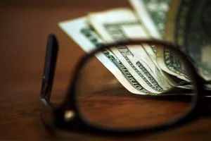 Some dollar bills in focus through a pair of glasses overturned on a desk.