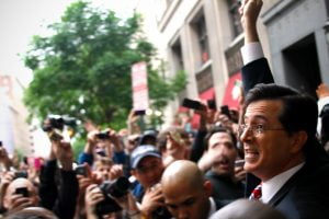 A triumphant Steven Colbert emerges from the FEC in Washington DC and holds money high above the crowd.
