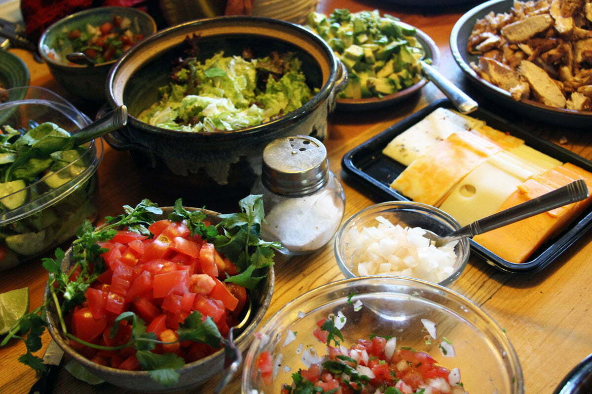 A wooden counter covered in usual taco fixings such as salsa, cheese, tomato, ground beer, grilled chicken, guacamole and lettuce.