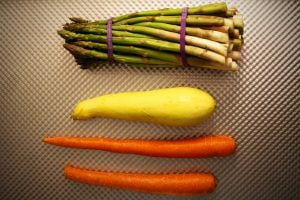 A bundle of asparagus, a single yellow squash and two carrots lie on a pan before cooking preparation begins.