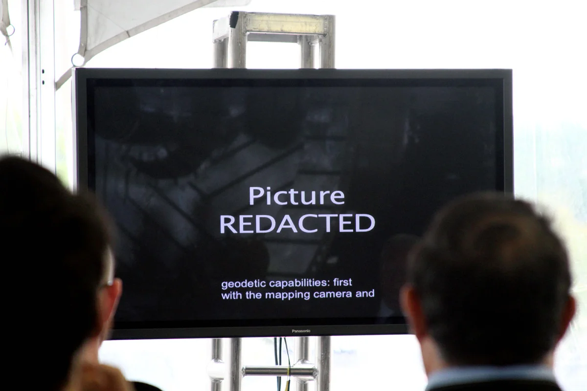 A video screen showing "Picture Redacted" as a crowd watches.