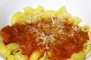 A white bowl of yellow pasta and some red sauce covered in white cheese.