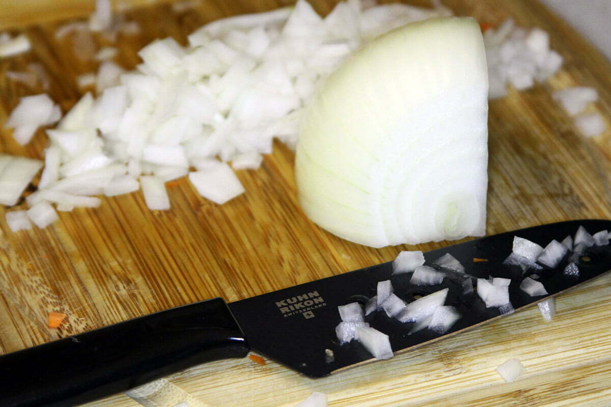 Half of an onion lies uncut on a wooden cutting board near a pile of diced onions and a black knife covered in more onion bits.