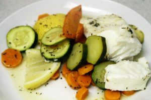 The finished halibut meal sits on a white plate surrounded by cooked veggies including lemons, zucchini and carrots.