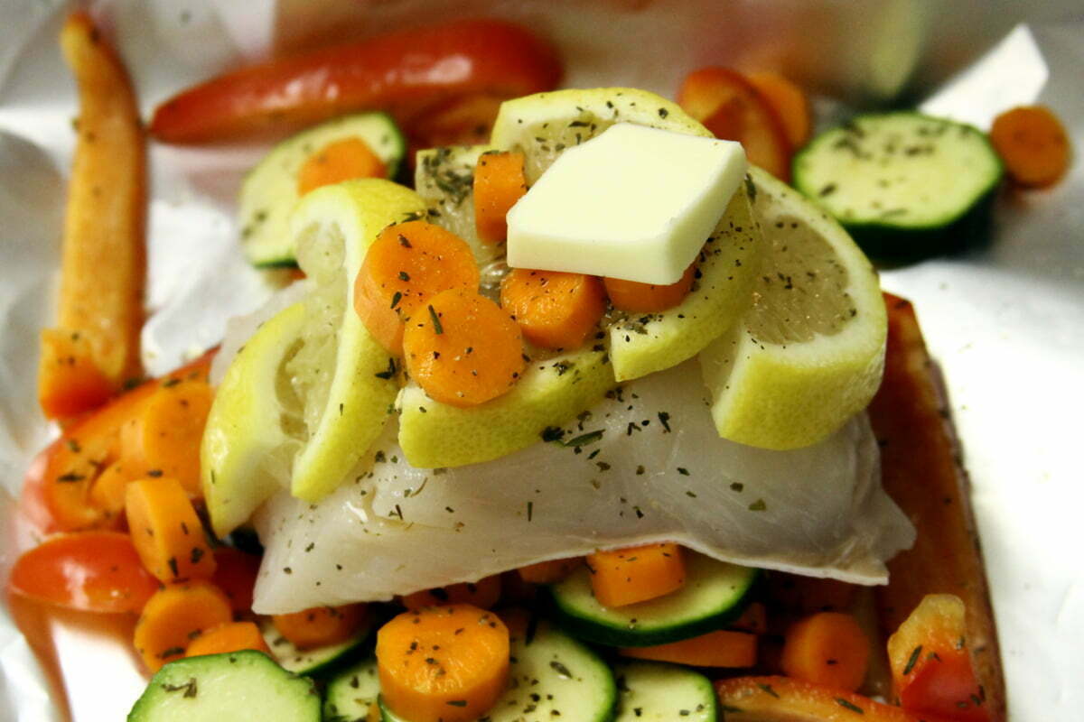 A filet of Halibut sits on some foil covered in lemon slices, carrots, cucumber and butter.