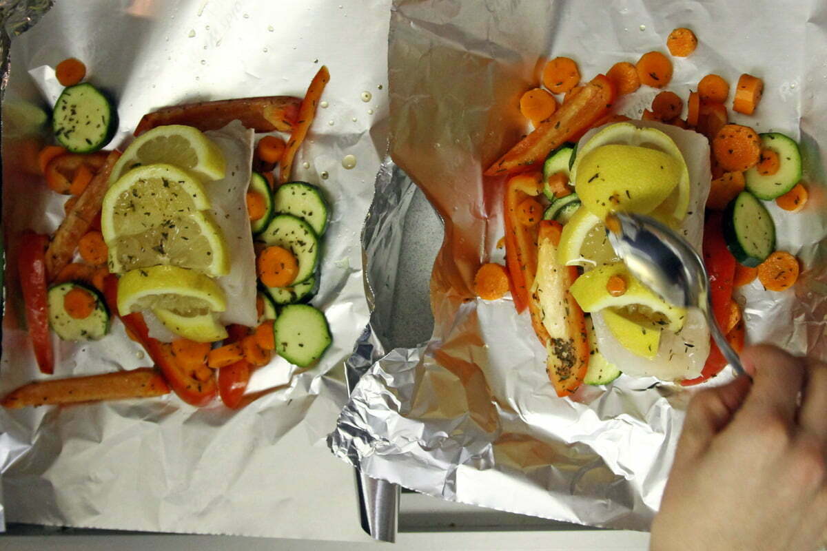 Two pieces of white Halibut fish sit on aluminum foil with colorful lemon wedges, zucchini slices, carrots and red pepper.