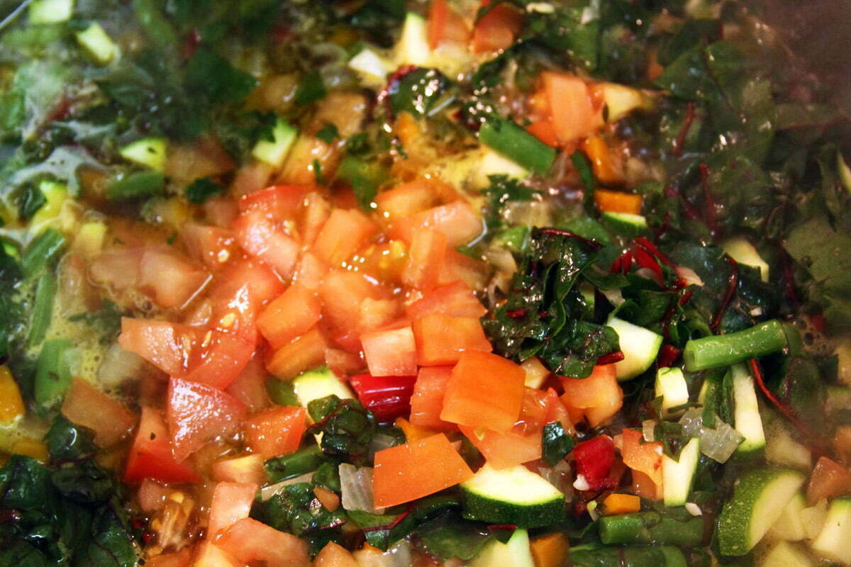 A pile of veggies in a big pot of soup - ingredients include cut tomatos, red chard, onion, garlic, carrot and green beans.