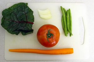 A carrot, tomato, string beans, bits of an onion and a leaf of a ruby chard sit on a white cutting board before being incorporated into a soup.