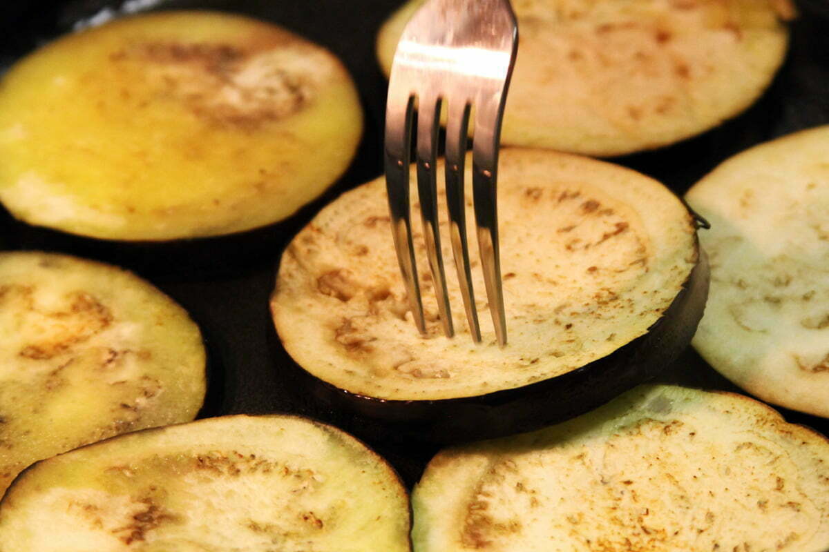 A fork turns slices of eggplant cook in a cast-iron pan.