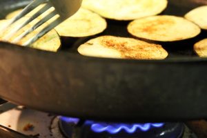 A cast iron skillet sits on a blue hot flame and cooks some pieces of eggplant.