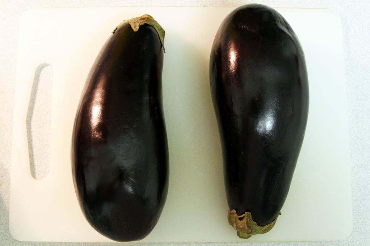 Two large dark purple eggplants sit side-by-side on a cutting board.