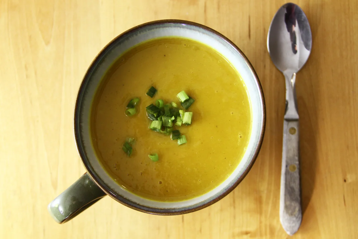 A large cup of butternut squash soup with a spoon on a wooden table, seen from above.