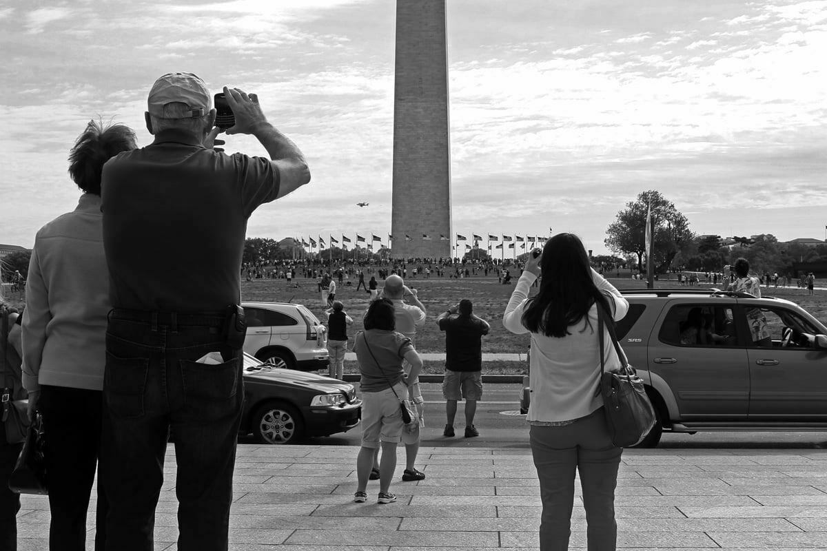 A crowd of people stop to photograph and watch the Space Shuttle Discovery flying by the Washington Monument.