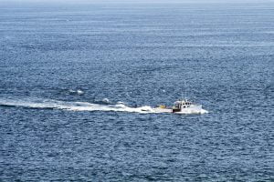 A Maine lobster boat traveling through blue choppy waters with gulls trailing behind near a lighthouse.