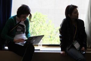 Two female TransparencyCamp attendees listen to a session and work on a laptop near a bright window.