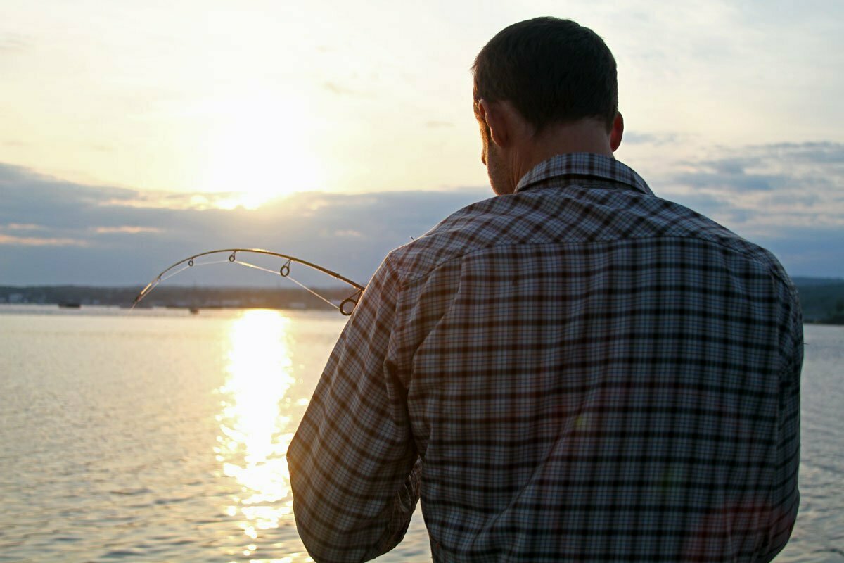 A man with his back to the camera focuses on his fishing line as the yellow sun sets over the water.