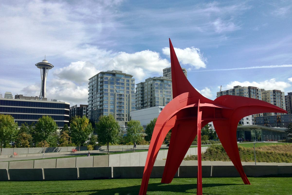 A large red metal sculpture called The Eagle by Alexander Calder is seen in the Olympic Sculpture Park in Seattle Washington with the Space Needle and skyline seen in the background.