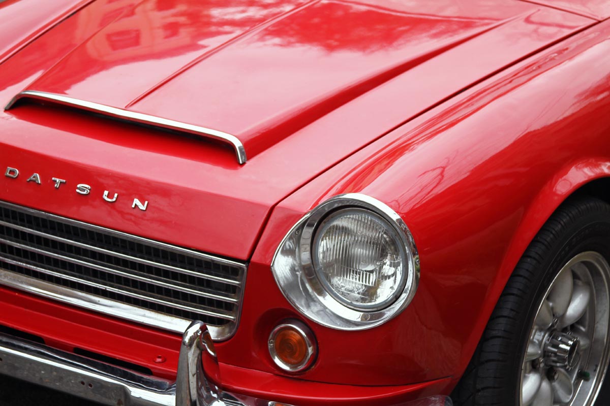 The front section of a red 1967 Datsun Roadster 1600 with the hood scoop, headlight, grill and panasport wheel visible.