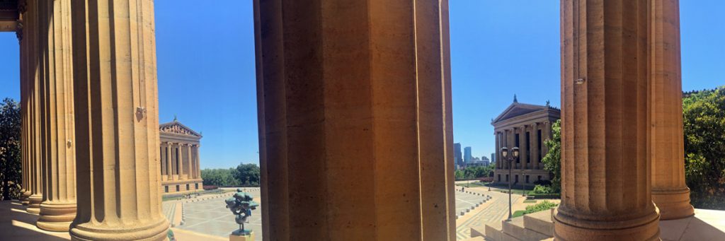 A panoramic photo taken from the steps at the east entrance to the Philadelphia Museum of Art. A series of columns break up the image with views of the open plaza and Greek buildings.