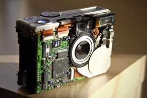 An old Canon PowerShot A50 being taken apart with the faceplate removed with the computer chip, lens and other internal components visible.