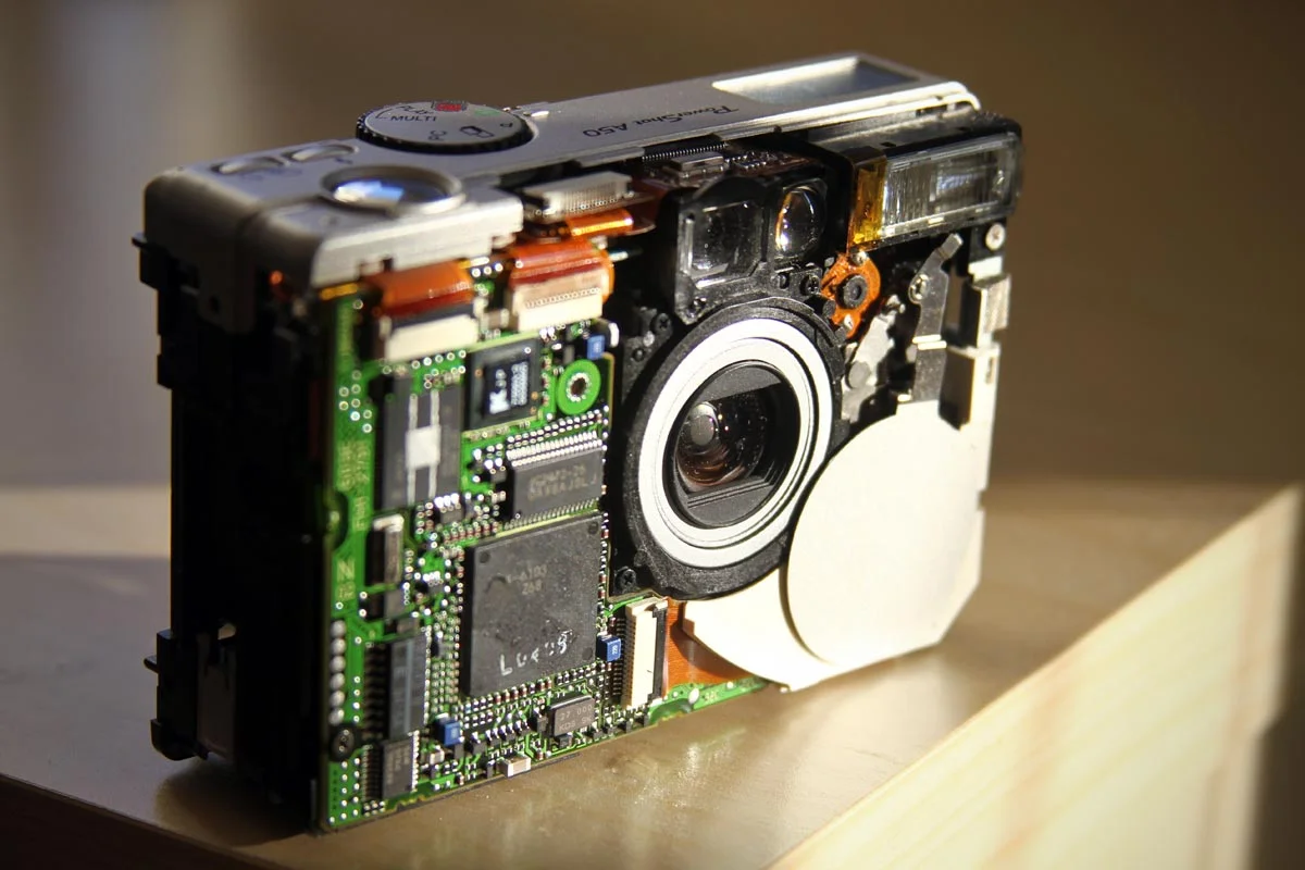 An old Canon PowerShot A50 being taken apart after the faceplate was removed with the computer chip, lens and other internal components visible.