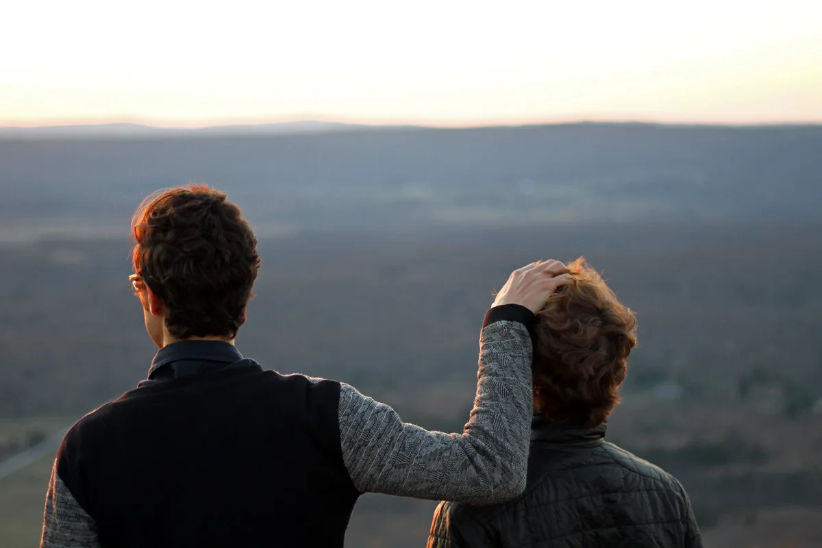 Two friends stand to watch the sun setting over a mountain with one running his hands through the hair of his friend.