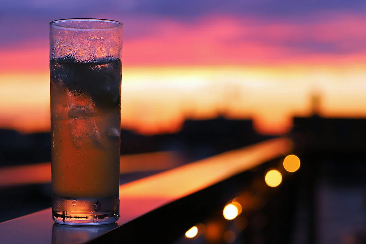 A photograph of a tall glass of gin and tonic with ice cubes sits on a ledge with a spectacular sunset in the background.