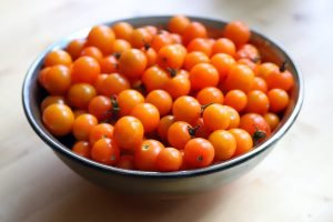 A bowl of bright red and orange cherry tomatoes sits in the sun on a wooden table.