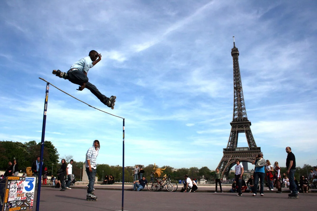 A Paris skater on rollerblades jumps over a high bar with onlookers and the Eiffel Tower in the background.