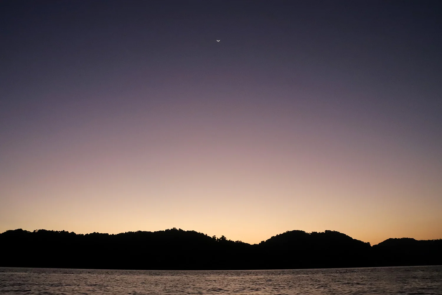 A purple and yellow sunset over an island in Mu Ko Surin National Park in Thailand with a crescent moon overhead.