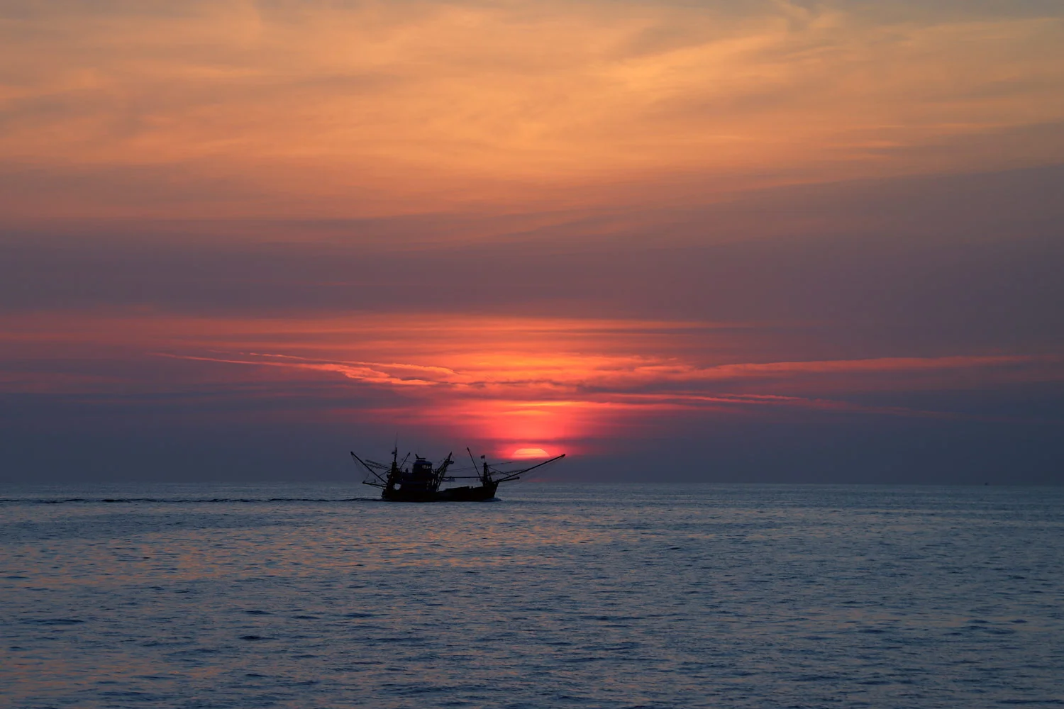 The sun sets on the ocean horizon next to a shrimper vessel rigged to start fishing off the coast of Thailand.
